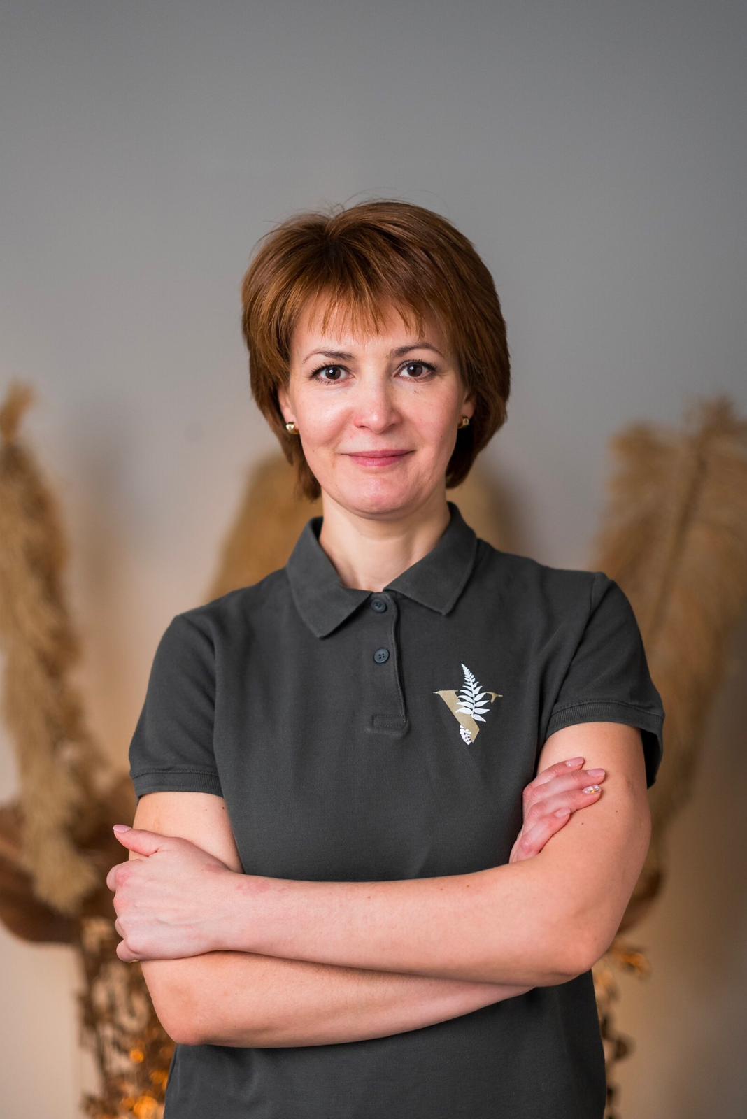 Eine lächelnde Frau in einem dunklen Polohemd mit Adler-Logo, verschränkte Arme, grauer Hintergrund.