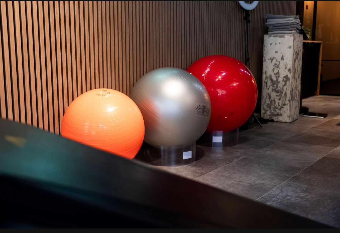 Drei Gymnastikbälle, orange, silber und rot, auf grauem Boden vor Holzwand im Innenbereich.