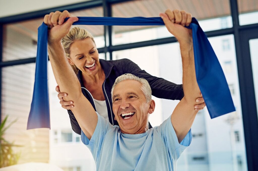 Sie hilft ihm, große Fortschritte zu erzielen. Aufnahme einer Physiotherapeutin, die einem älteren Patienten in ihrer Praxis beim Dehnen mit einem Stretchband hilft.