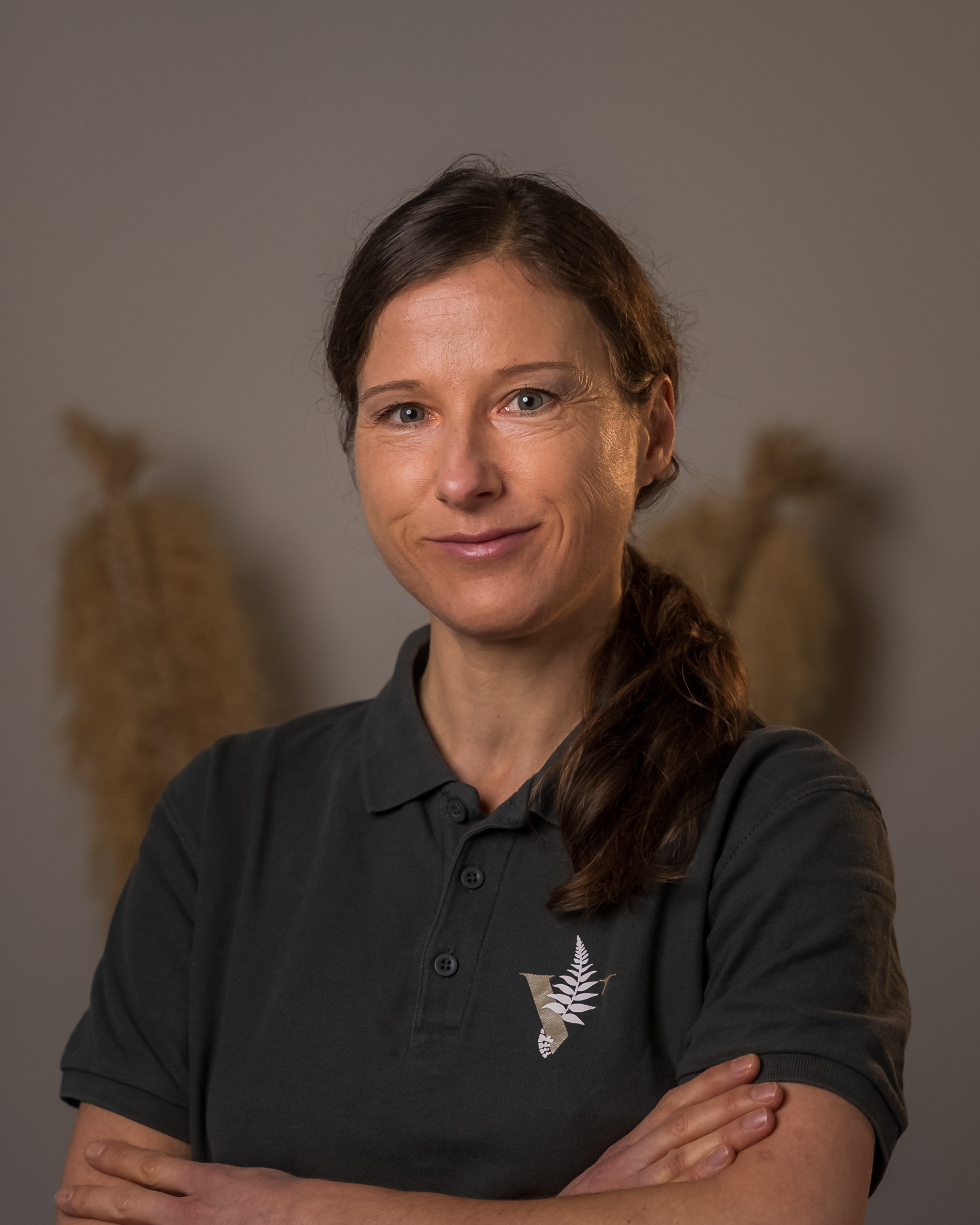 Eine lächelnde Frau mit braunen Haaren im Zopf trägt ein dunkles Poloshirt vor beiger Dekoration