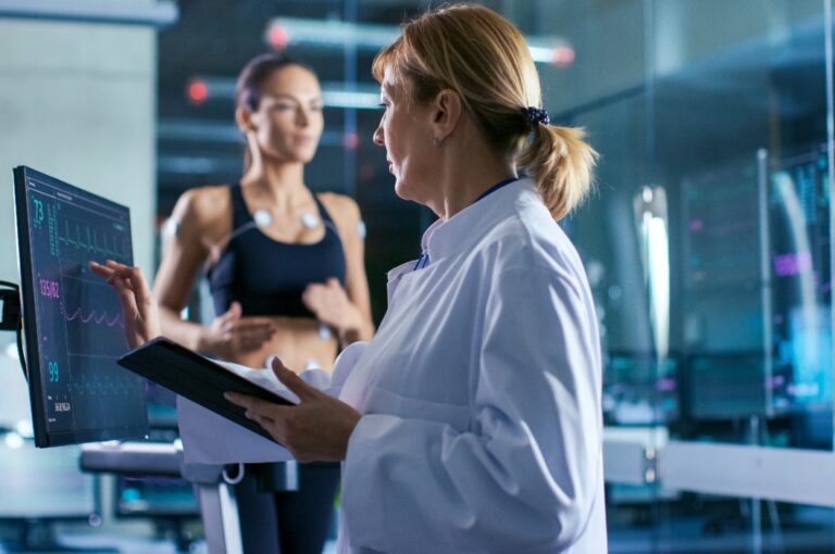 Ein Sportwissenschaftler beobachtet eine Anzeige, die den EKG-Status anzeigt, während im Hintergrund eine Sportlerin auf einem Laufband läuft