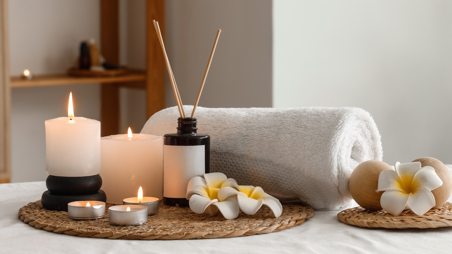 Ein Wellness- und Entspannungstherapie-Ambiente mit brennenden Kerzen, einem Aromadiffusor, einem aufgerollten Handtuch und Plumeria-Blüten auf einer Massageliege